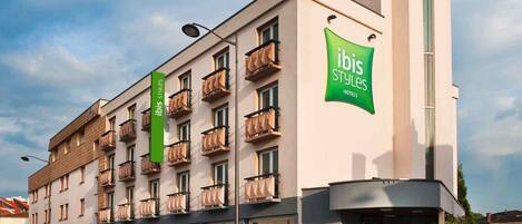 Un hotel de varios pisos con balcones, un letrero verde de 'ibis Styles Hotels' y una fachada moderna.