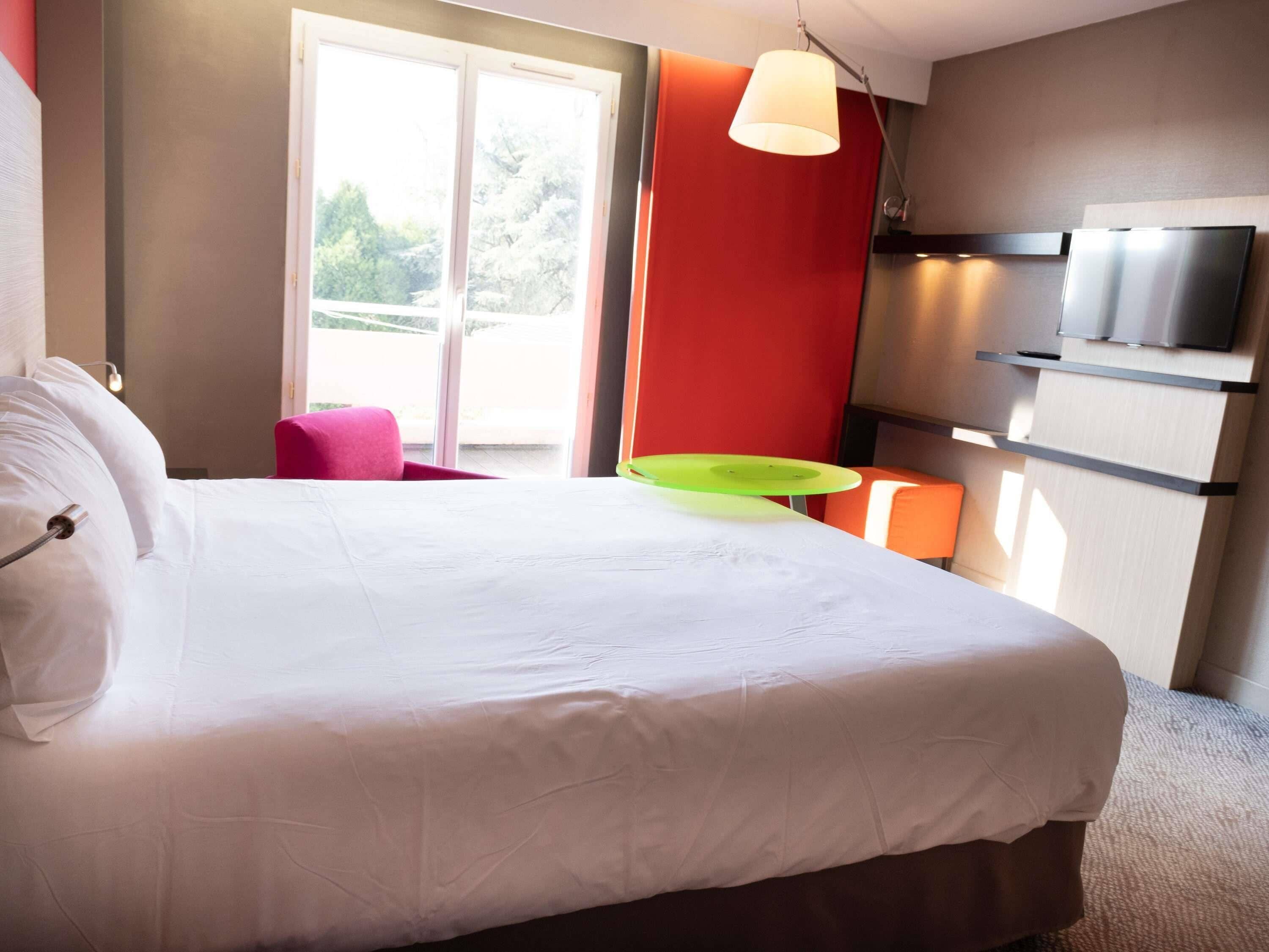 A hotel room with a bed, a green chair, a red wall, and a TV mounted on the wall.