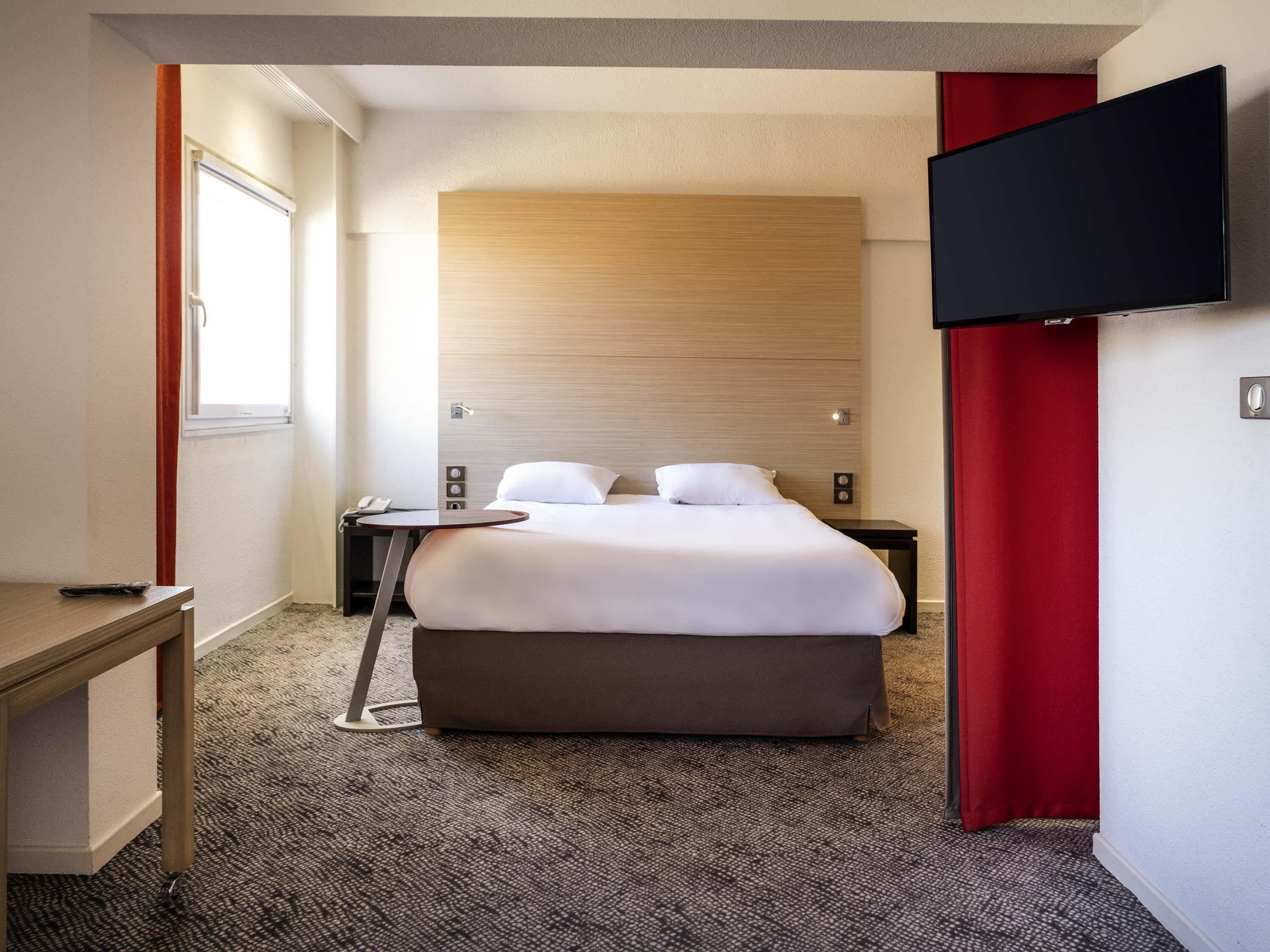 A hotel room with a bed, a TV, a desk, and a window.
