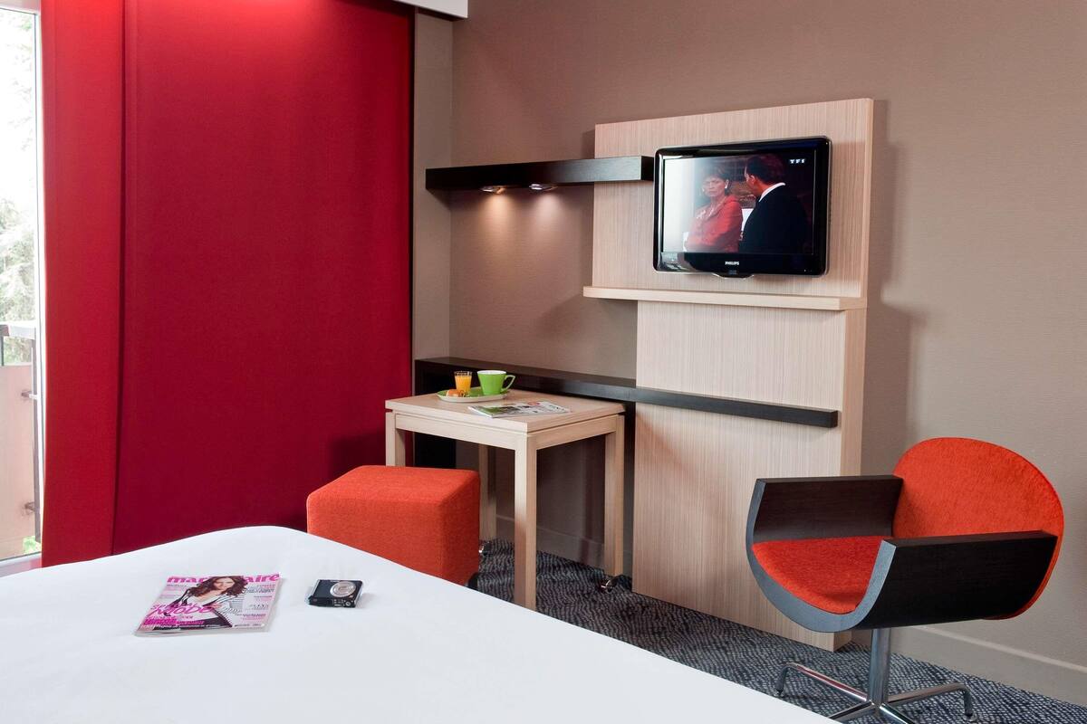 Habitación de hotel con cama, televisor montado en la pared, una mesita con una taza y una revista, y una silla roja.