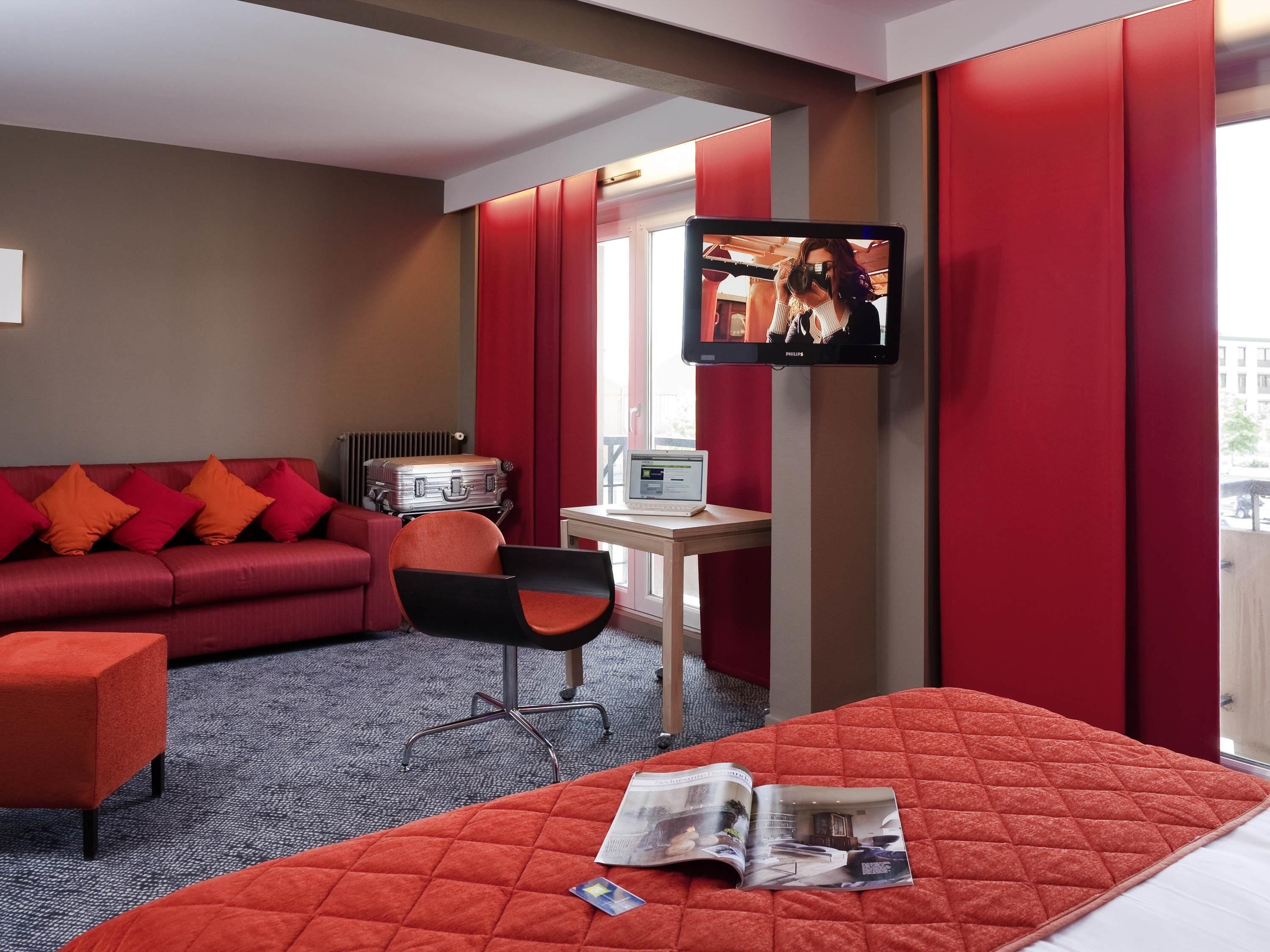 A hotel room with a red sofa, a desk with a laptop, a television, and a bed with a magazine.