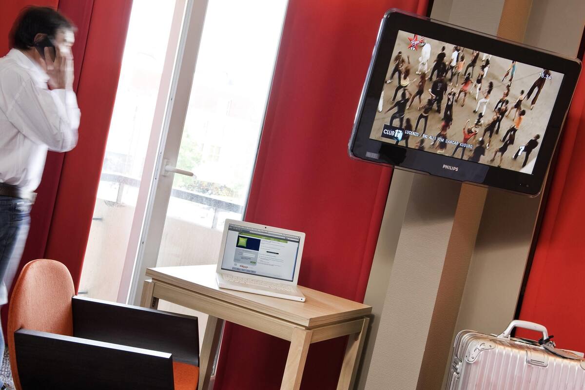 Habitación de hotel con una televisión mostrando un programa de baile, una computadora portátil en un escritorio, una silla y una maleta.
