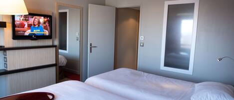 A hotel room with a TV on the wall, a bed with white linens, a bedside table, and a door leading to another room.