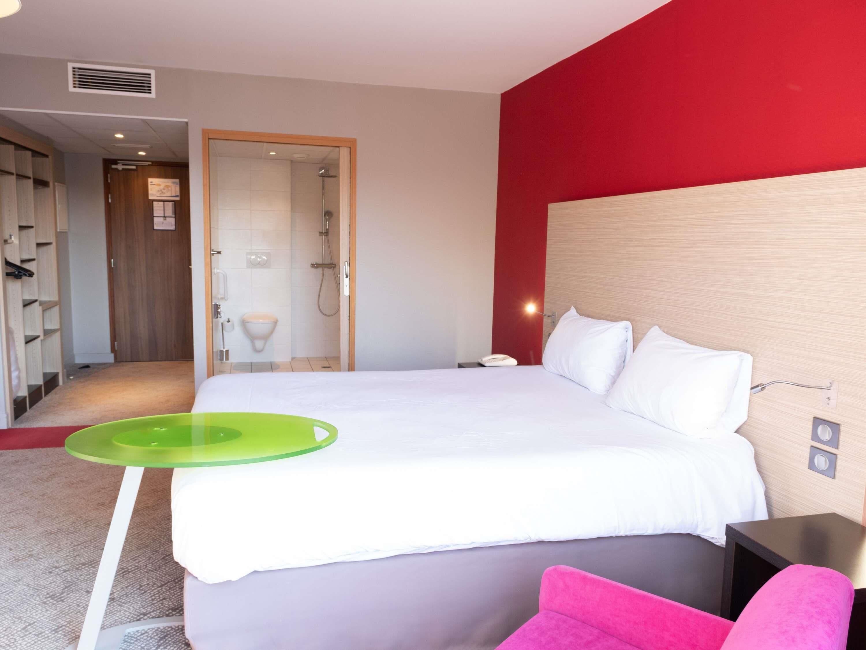 A hotel room with a bed, a green tray, a pink chair, a wooden headboard, and a red accent wall.