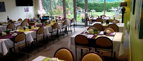 A dining area with multiple tables and chairs, large windows with a view of greenery, and a flat-screen TV mounted on the wall.
