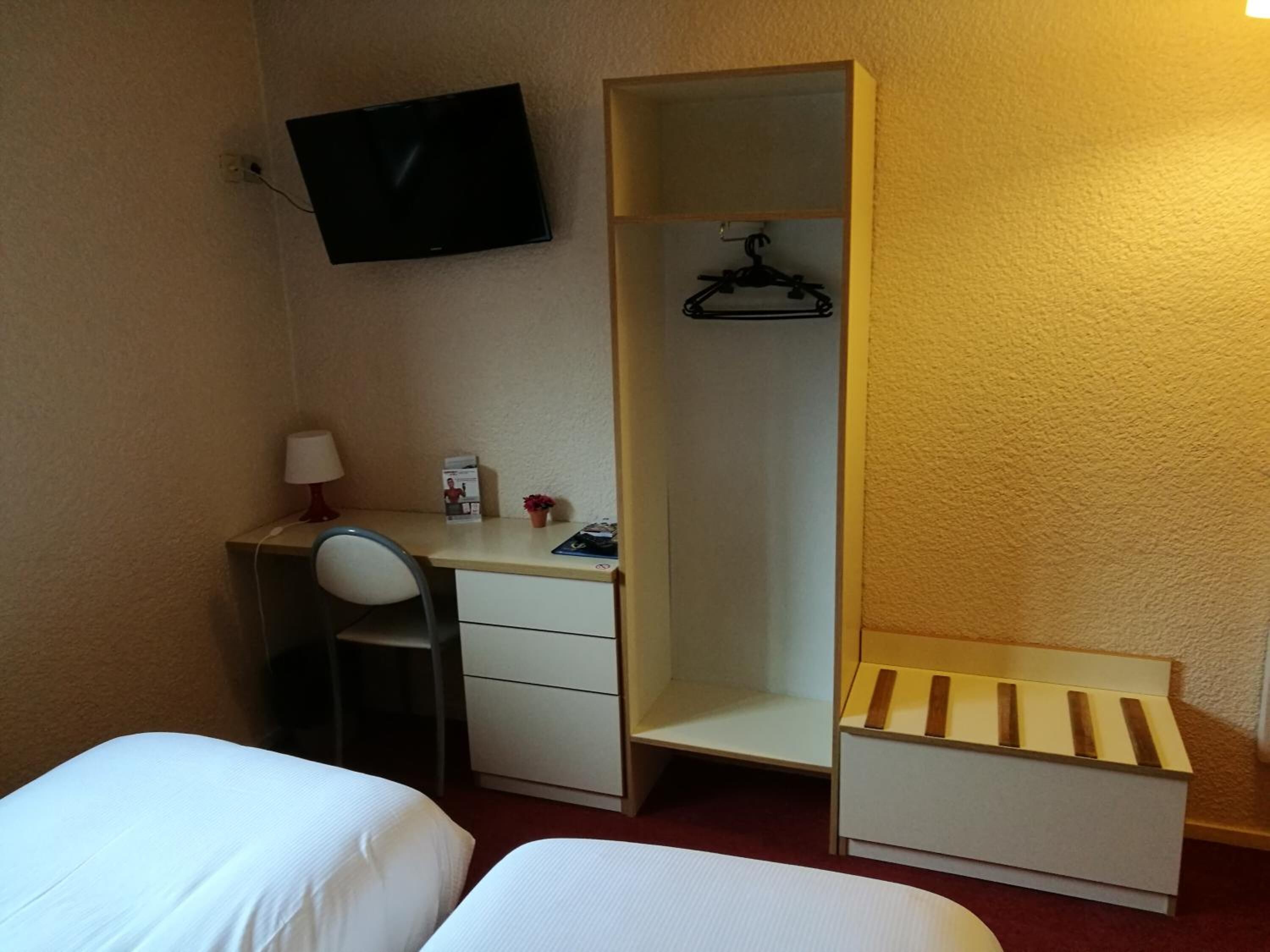 A small hotel room with a desk, two beds, a wardrobe, a TV, and a small shelf.