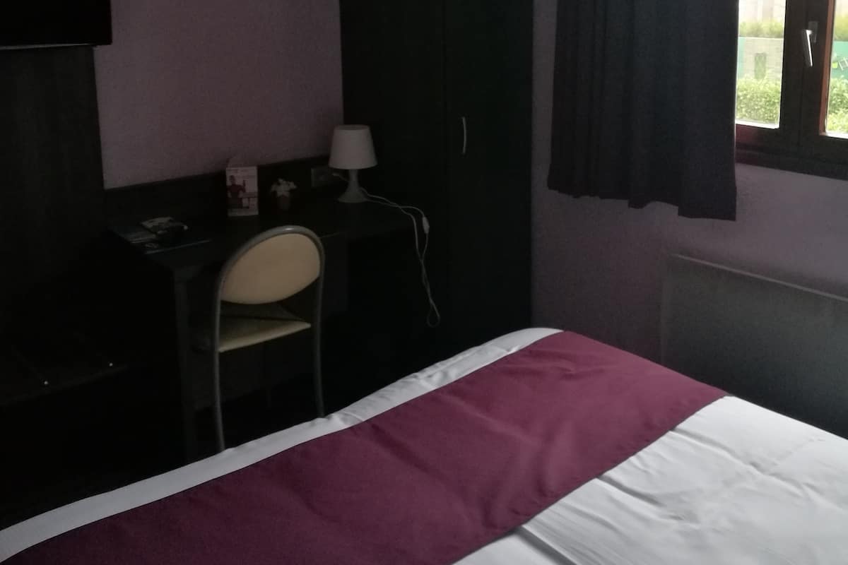 A hotel room with a bed, a desk with a chair, a wardrobe, and a window with curtains.