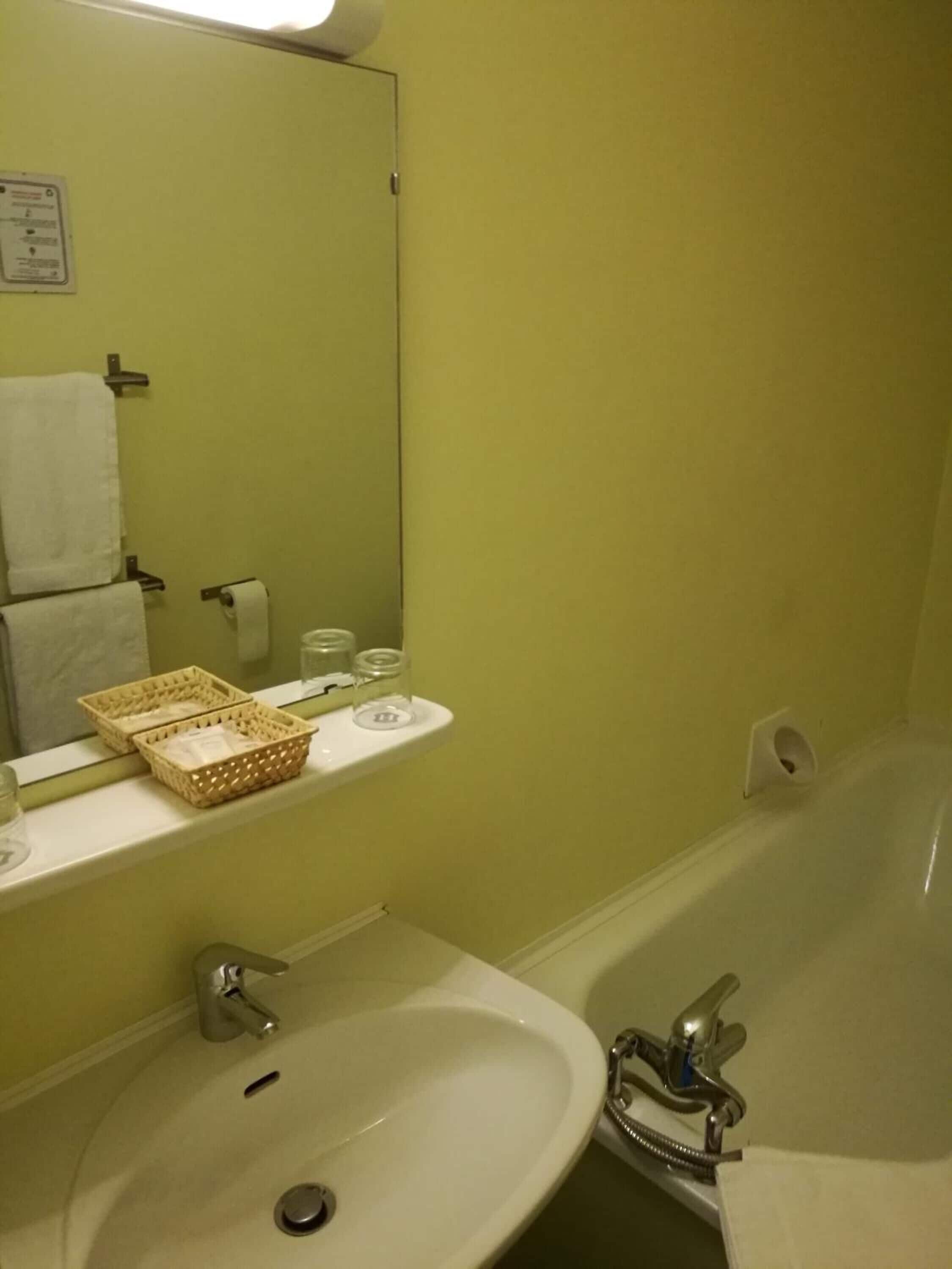 Een badkamer met een wastafel, spiegel en badkuip. Op de wastafel zitten twee manden, er hangt een handdoek aan de muur en staat een toiletpapierhouder.