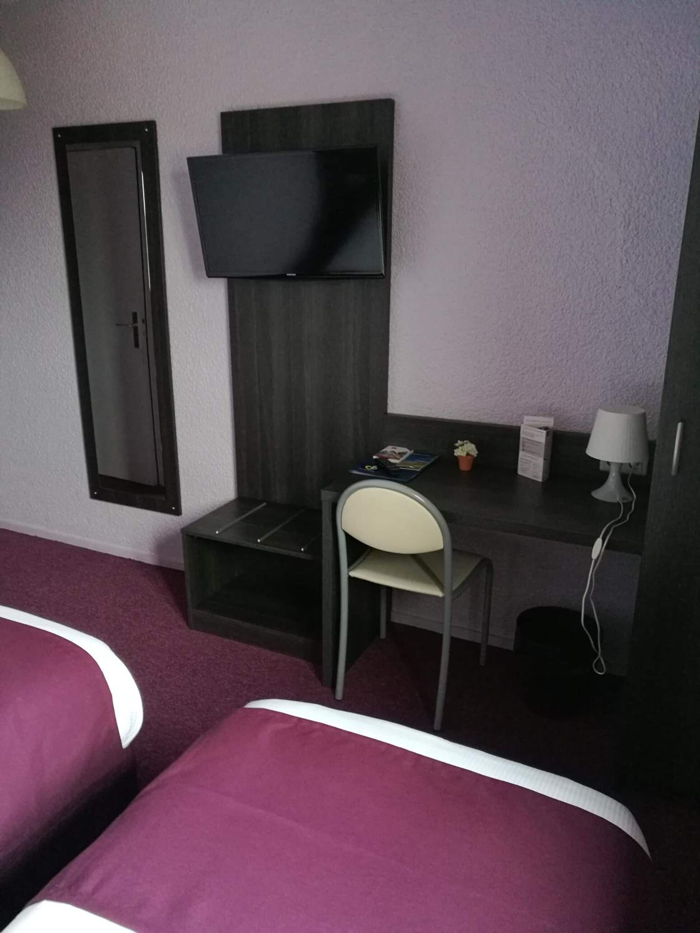 A hotel room with two beds, a desk with a chair, a flat-screen TV mounted on the wall, and a small table with a lamp.