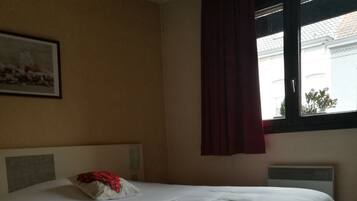 A single-bed room with a headboard, a window with a curtain, a radiator, and a framed picture on the wall.