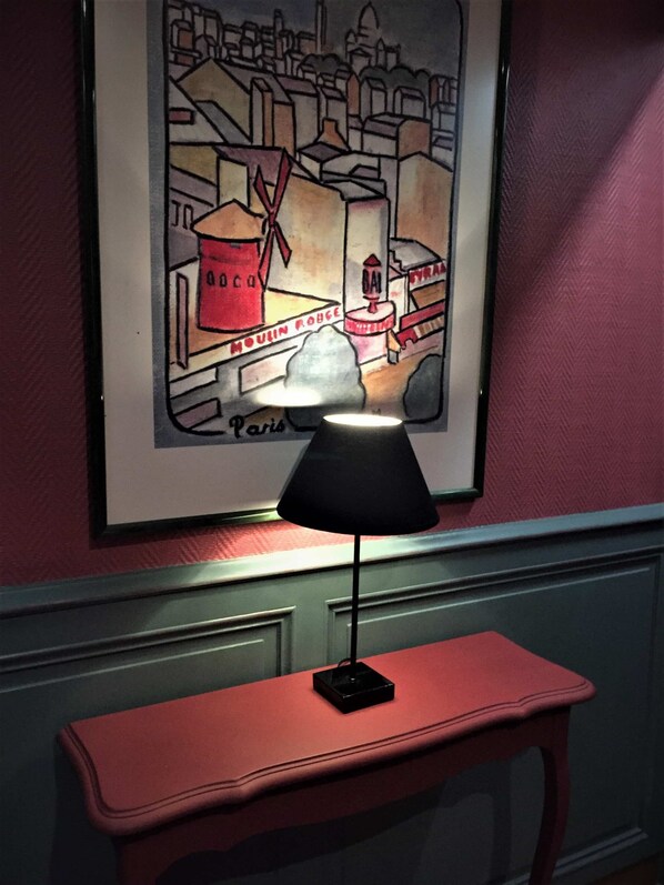 Una obra de arte con un paisaje urbano enmarcado en una pared, una lámpara de mesa negra y una mesa roja.