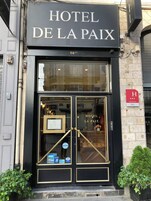 Entrada de un hotel con fachada negra, puertas dobles y un cartel que dice 'HOTEL DE LA PAIX'.