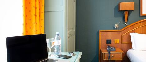 A hotel room with a bed, a bedside table with a lamp, a laptop, a smartphone, and a water bottle.