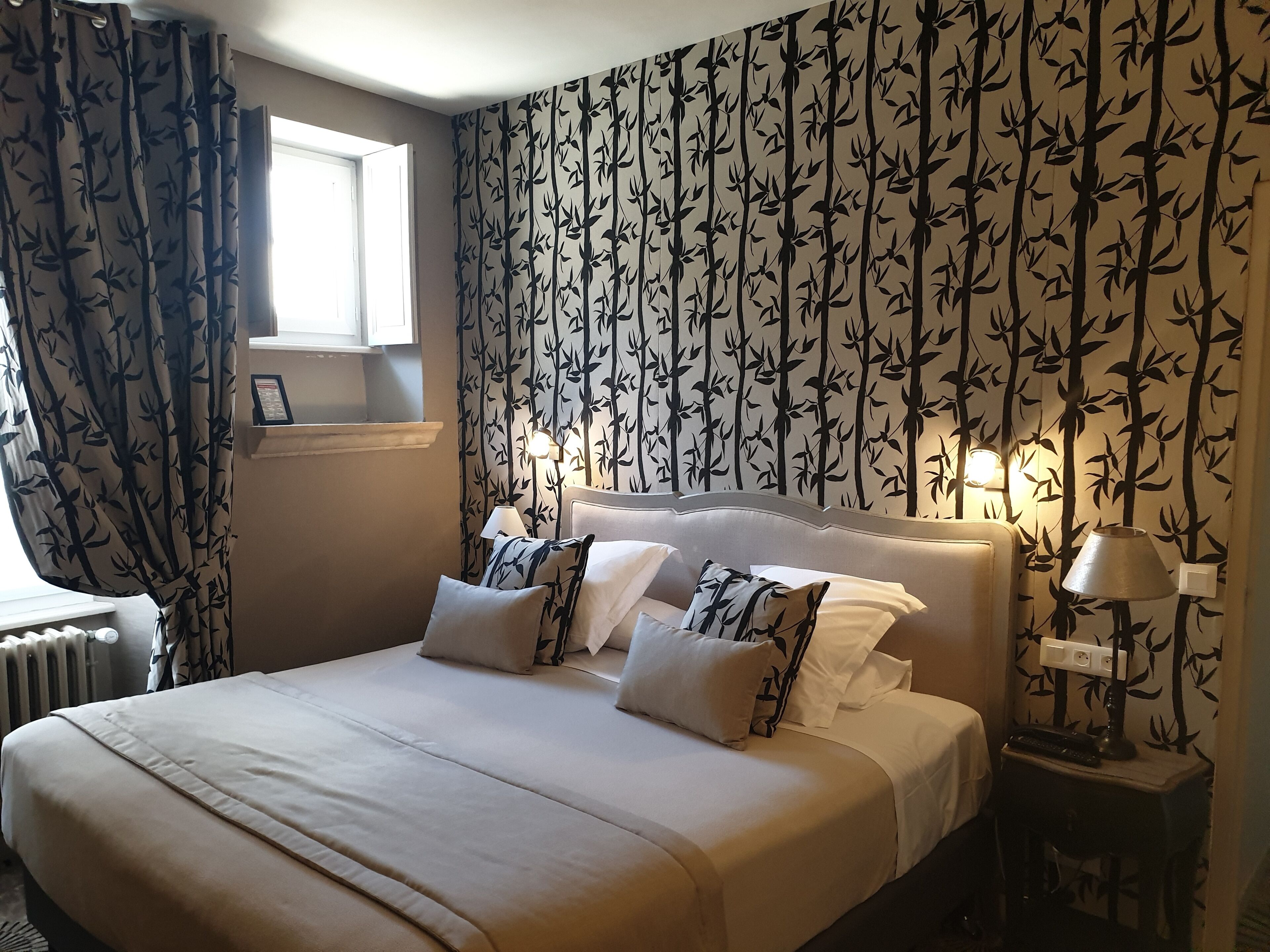 A neatly made bed with white linens and patterned pillows, a bedside table with a lamp, and a wall with a bamboo leaf pattern.