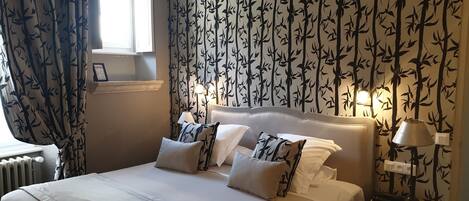 Una cama bien hecha con sábanas blancas y almohadas estampadas, una mesita de noche con una lámpara, y una pared con un diseño de hojas de bambú.