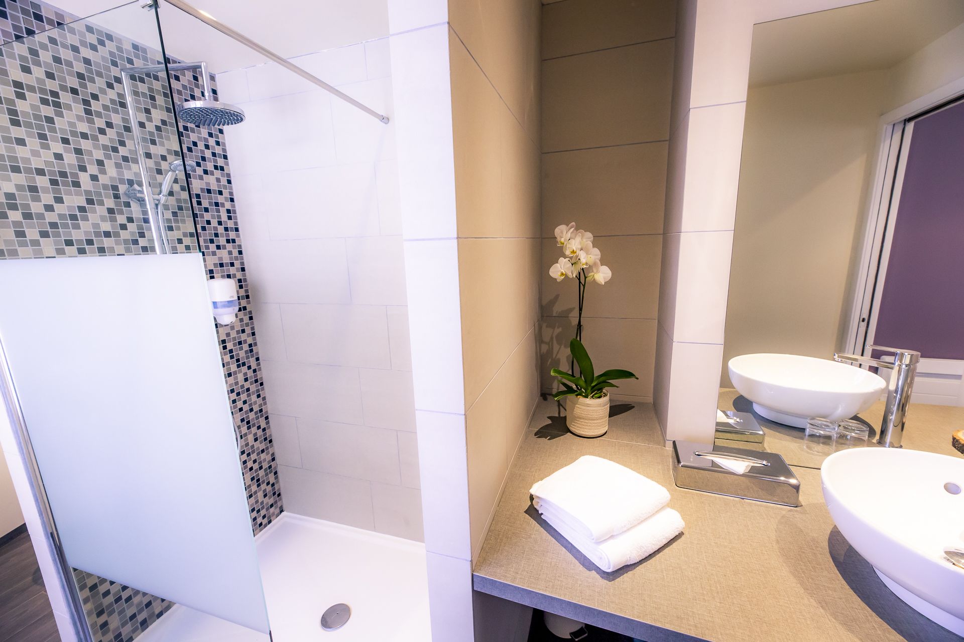 Un baño moderno con ducha, dos lavamanos, un espejo y una orquídea en maceta.