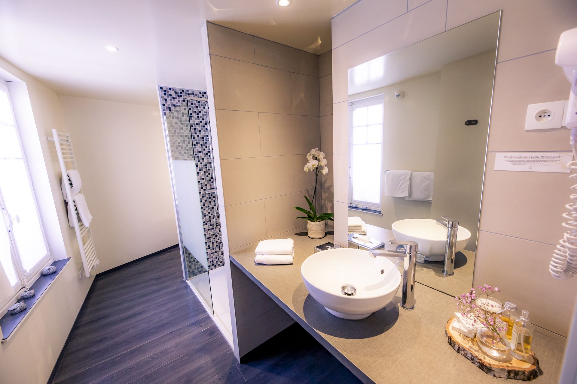 Une salle de bain moderne avec un grand miroir, un lavabo rond, une cabine de douche et une orchidée en pot.