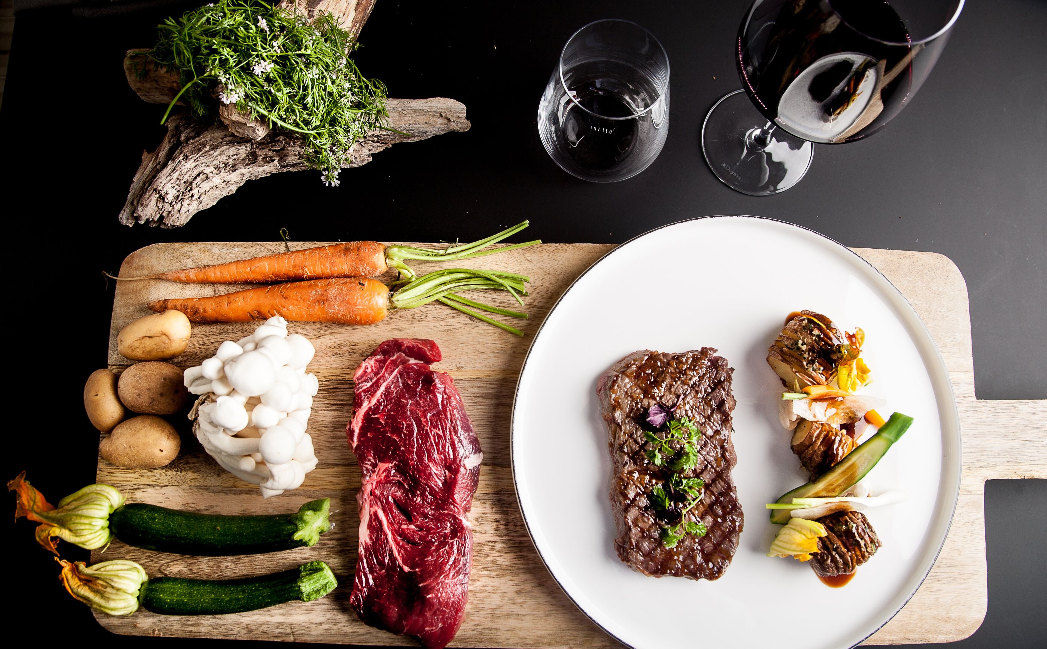 Una tabla de madera con varios vegetales y un bistec, un plato con bistec y vegetales, y dos copas de vino tinto sobre una superficie oscura.