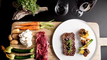 Una tabla de madera con varios vegetales y un bistec, un plato con bistec y vegetales, y dos copas de vino tinto sobre una superficie oscura.