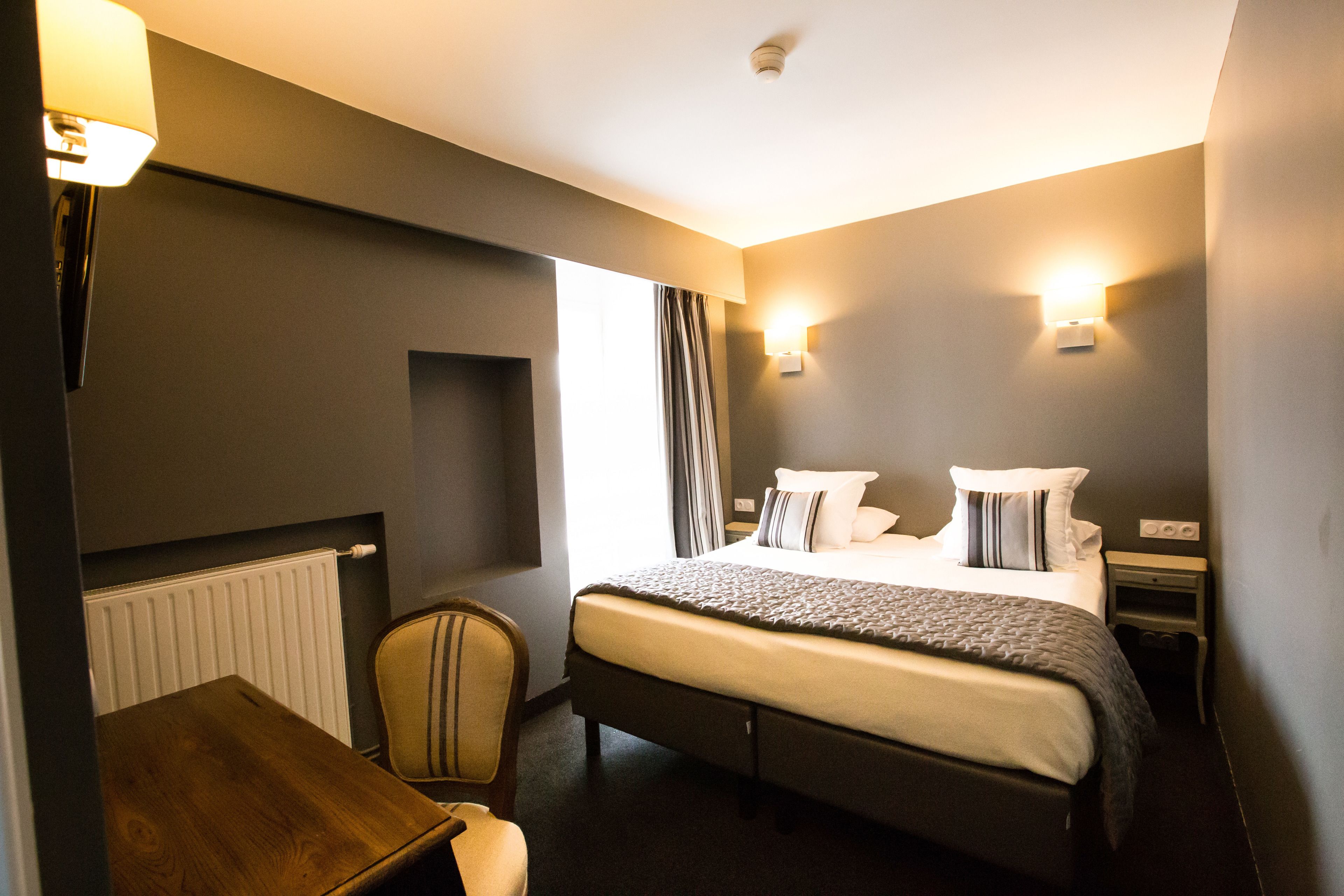 A hotel room with a bed, bedside table, chair, lamp, and wall-mounted light.
