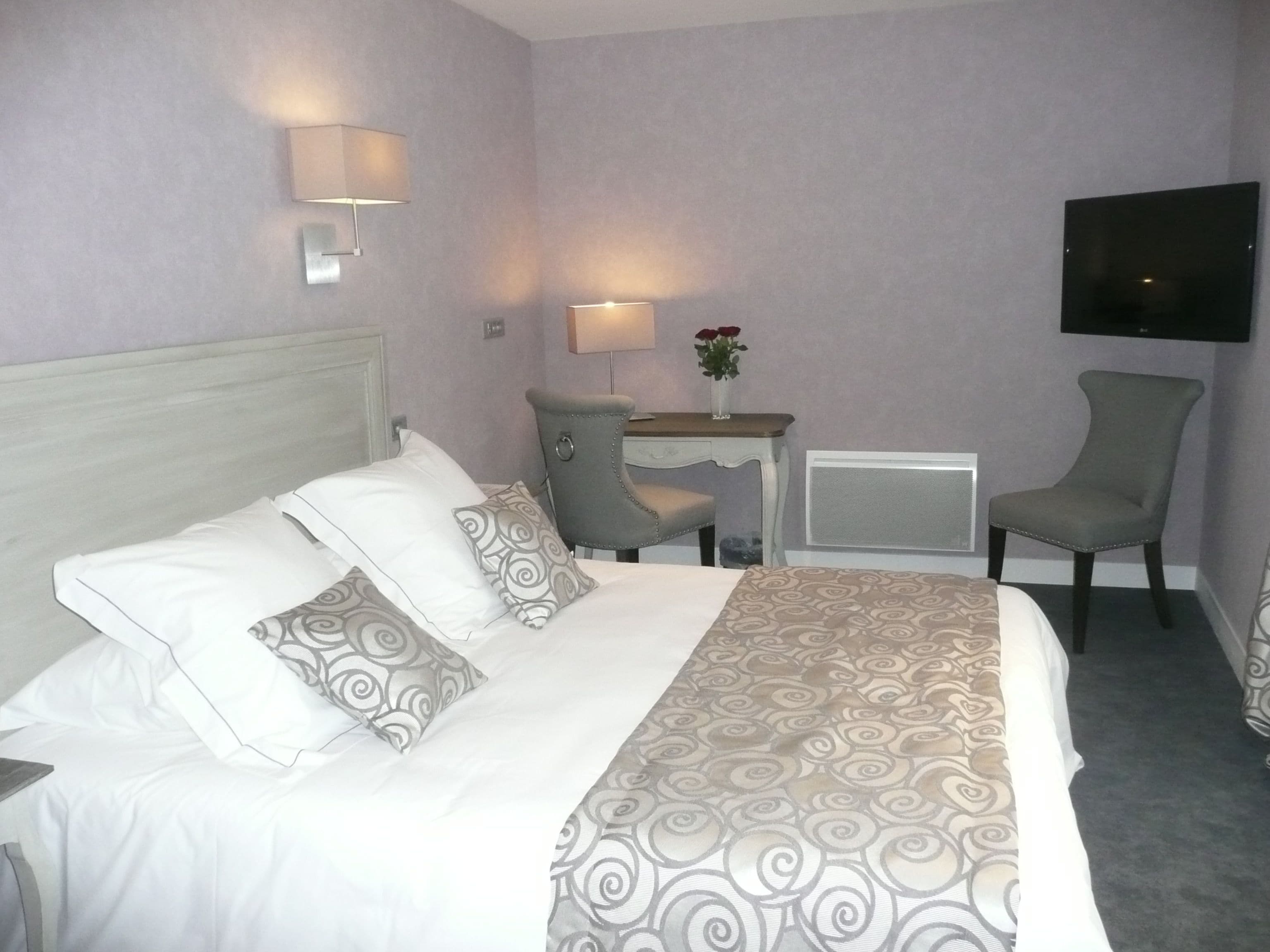 A hotel room with a bed, two chairs, a desk, a TV, and a vase with flowers.