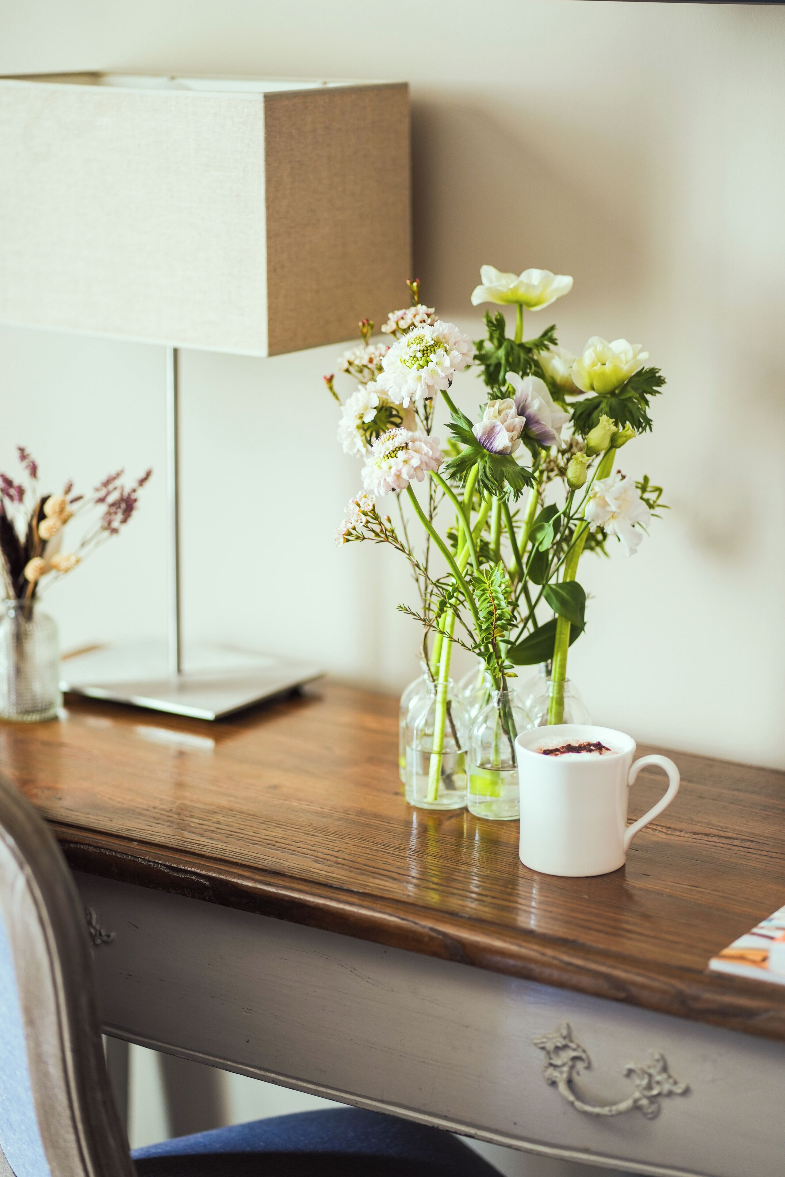Un escritorio de madera con una taza blanca, un florero de vidrio con flores y una lámpara.