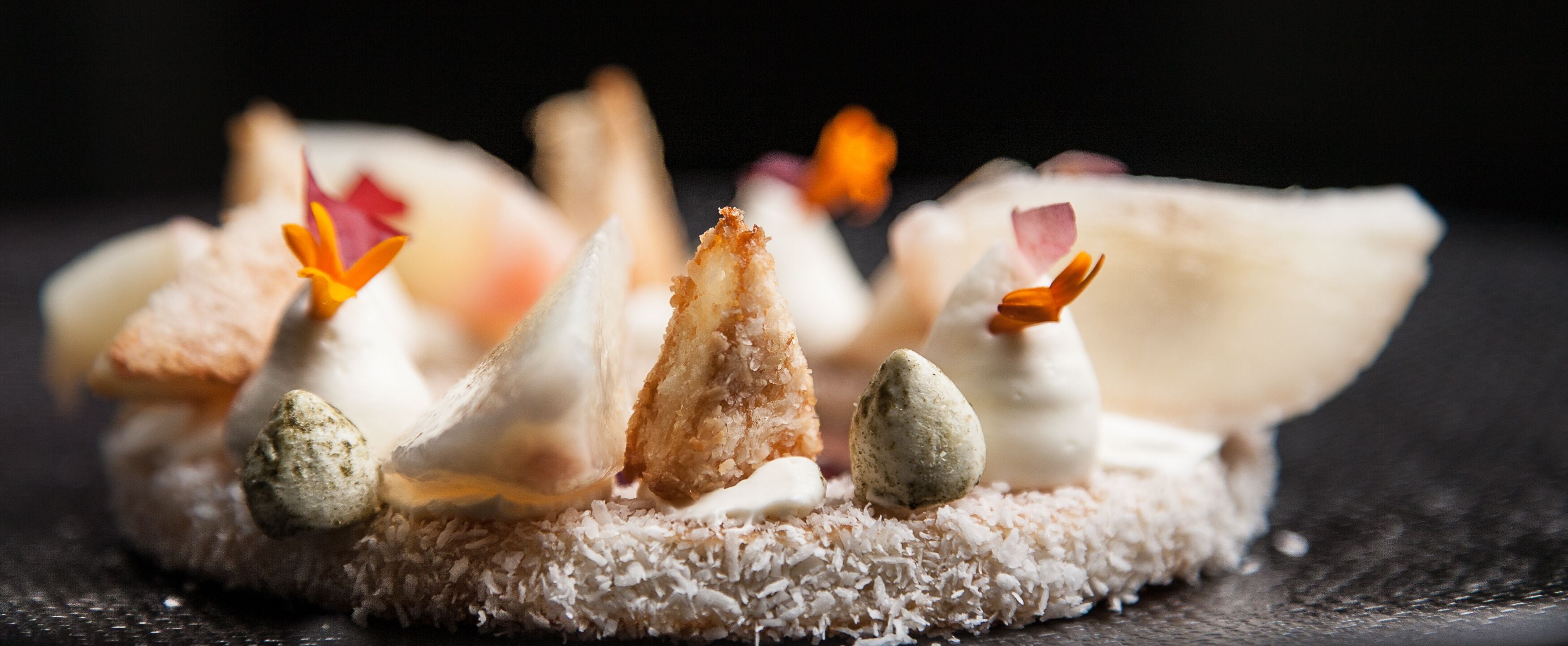 A dessert with various components including coconut shavings, a crispy element, and small edible flowers.