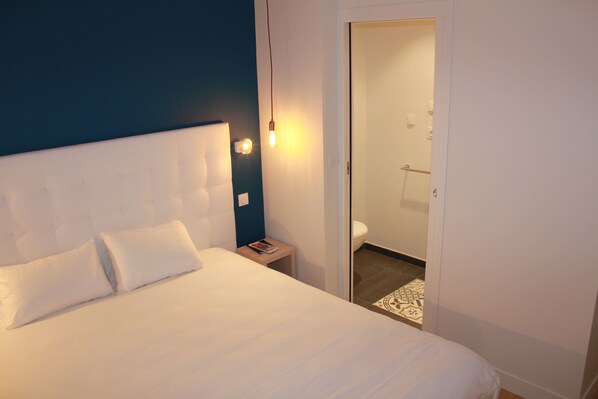 A hotel room with a large bed, a bedside table, a nightstand, and a door leading to a bathroom. - Hôtel La Fontaine Caen Centre (Caen)