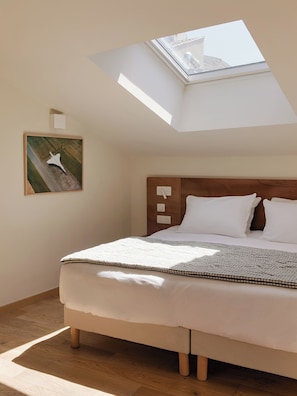 Une chambre avec une lucarne, un lit avec une literie blanche, une tête de lit en bois et un tableau accroché au mur.