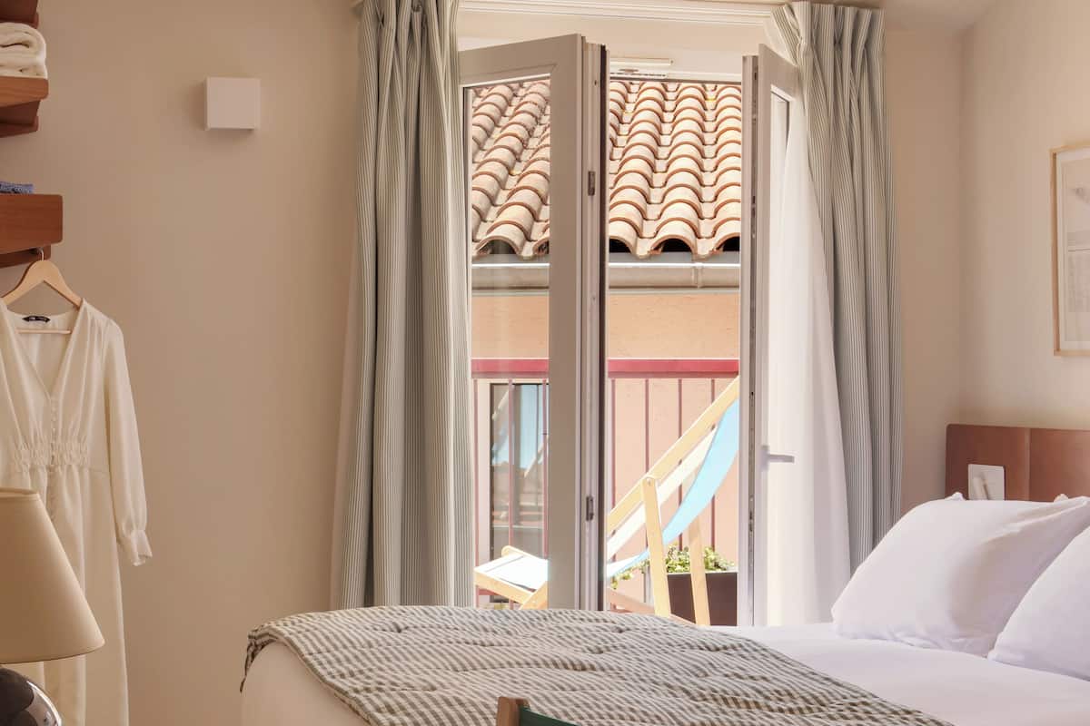 Una habitación de hotel con cama, escritorio, silla, ventana con cortinas y vista a un edificio con tejado de tejas.