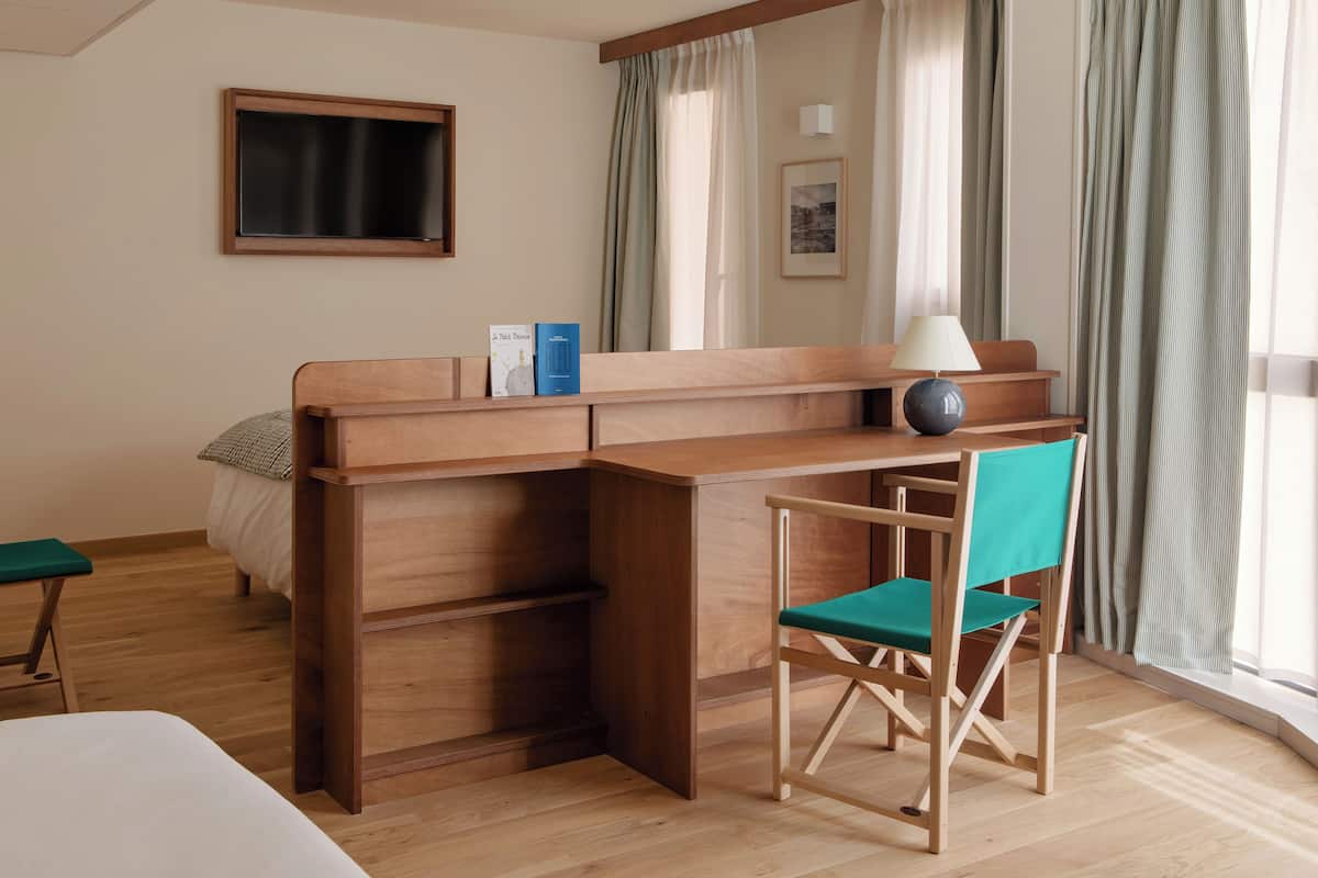 Habitación de hotel con un escritorio de madera, una silla verde, una cama, una mesita y una ventana con cortinas.