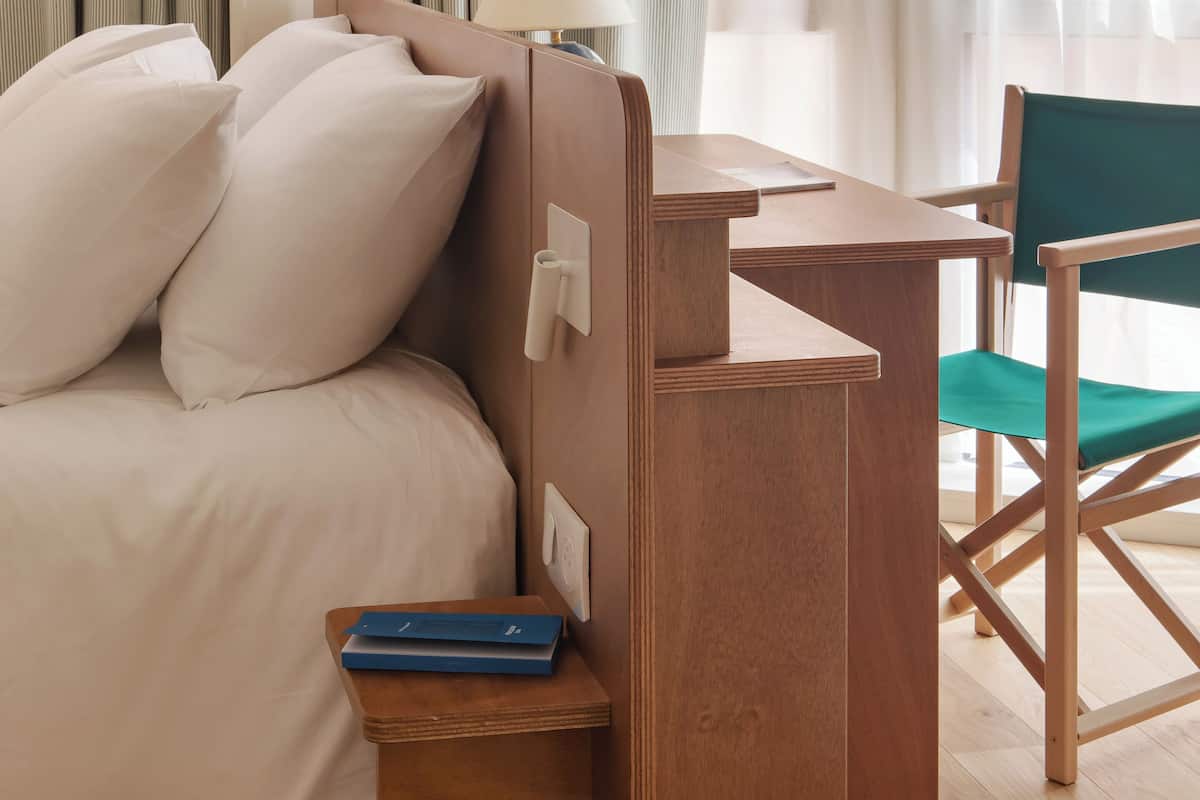 Un dormitorio con cama, mesita de noche, lámpara, silla y un pequeño estante de madera con libros.