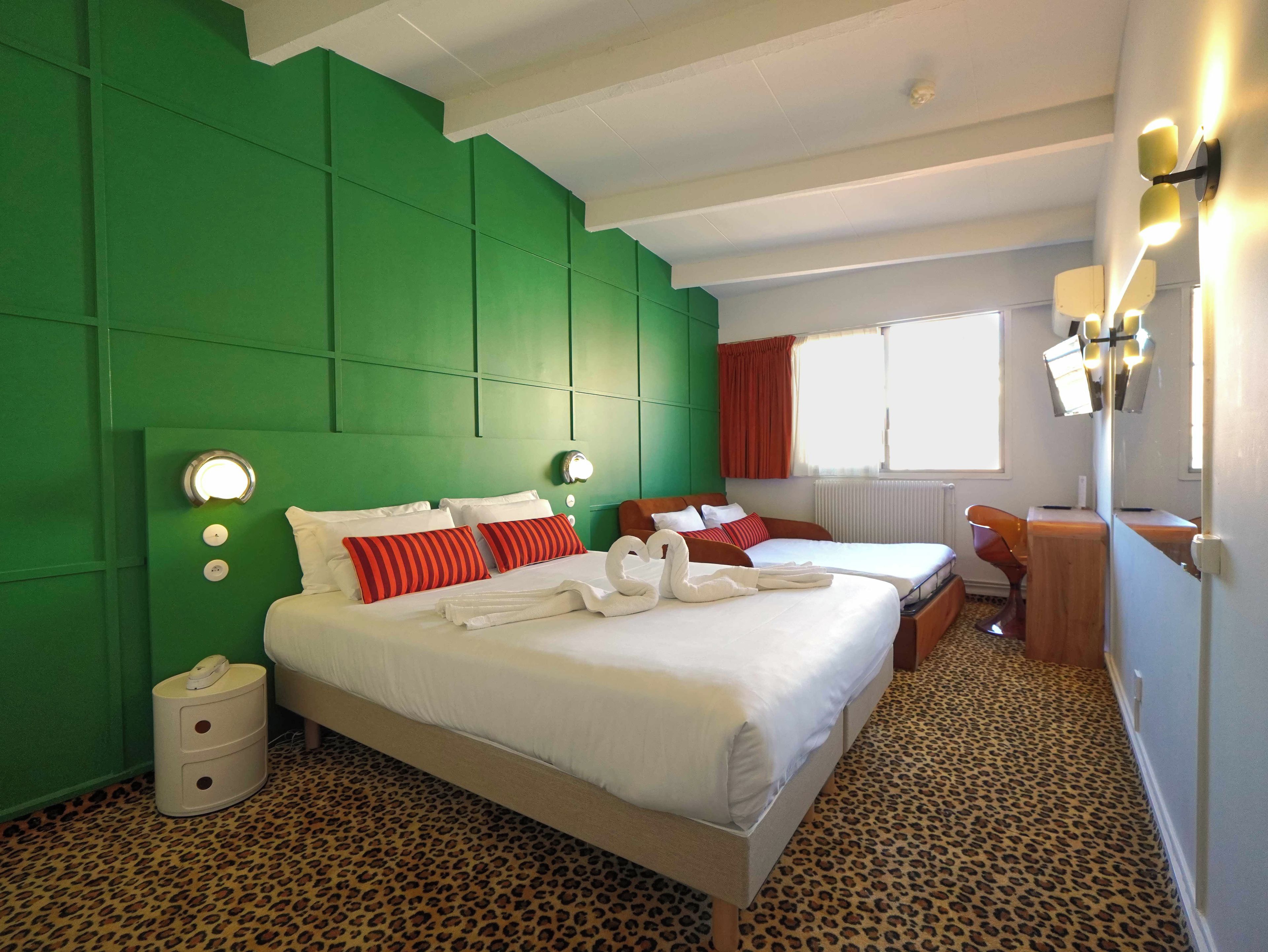 Une chambre d’hôtel avec un mur d’accent vert, un lit avec du linge de lit blanc, une moquette à motifs léopard et un bureau avec une chaise.
