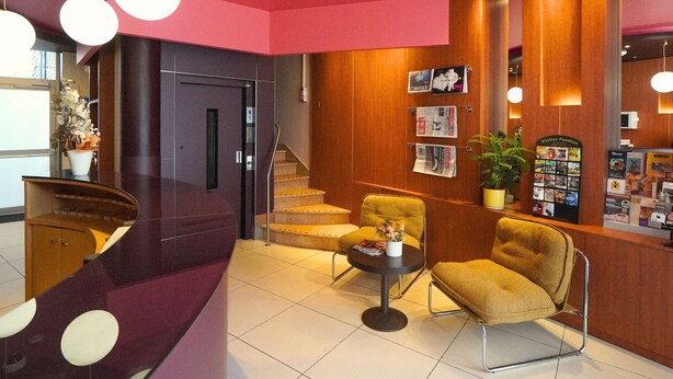 A reception area with a desk, two chairs, a coffee table, a staircase, and a bulletin board with various items. - Hôtel Le Moderne (Toulouse)