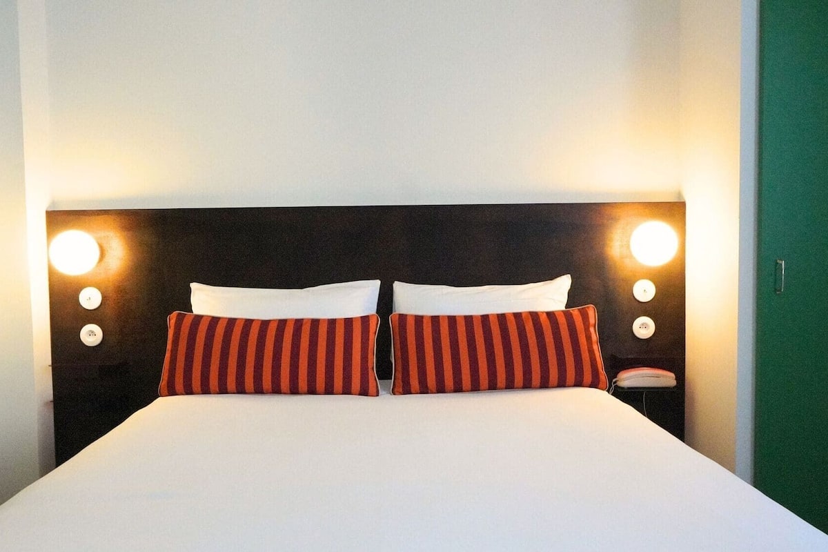 Un lit avec du linge de lit blanc, deux coussins à rayures rouges et noires et deux appliques murales rondes, une de chaque côté.