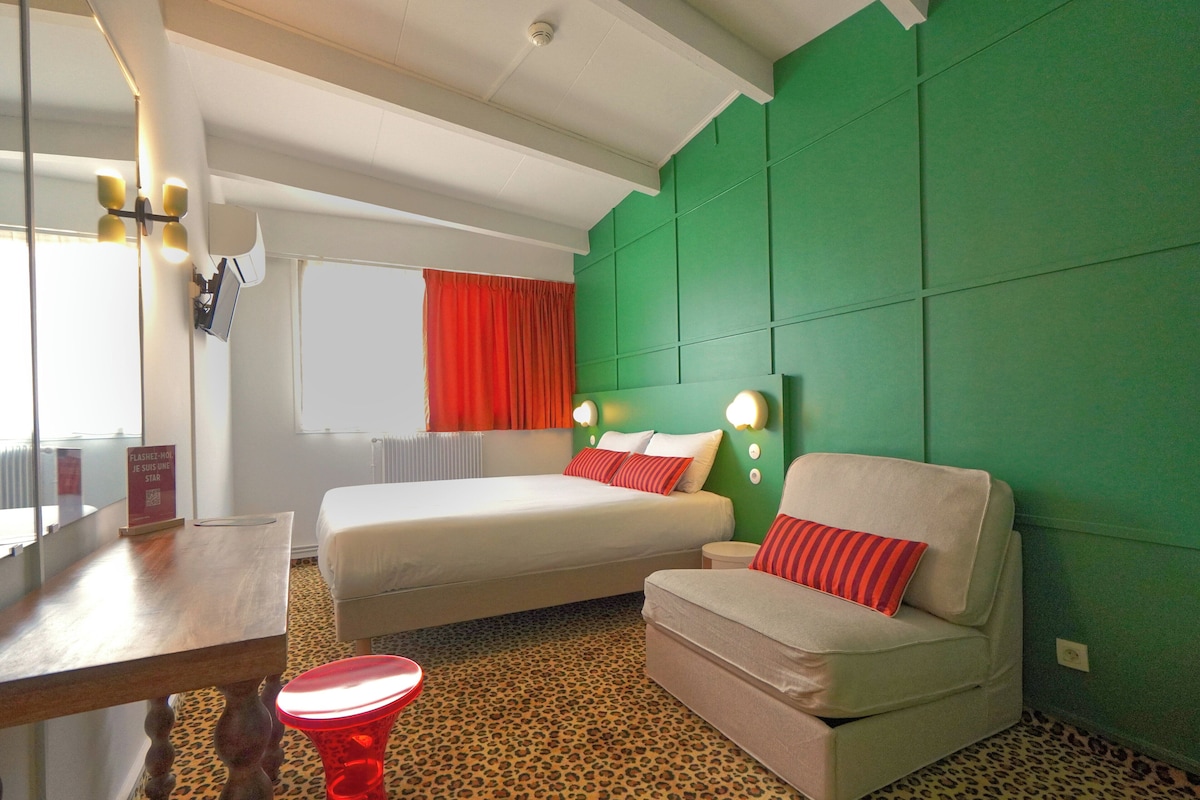 Habitación de hotel con dos camas, una pared de acento verde, alfombra de estampado de leopardo y una lámpara de mesa roja.
