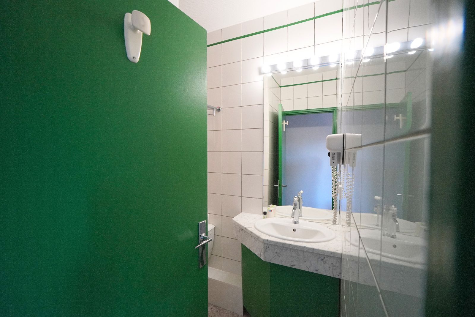 Un baño con puerta verde, azulejos blancos, lavamanos con espejo y secador de pelo.