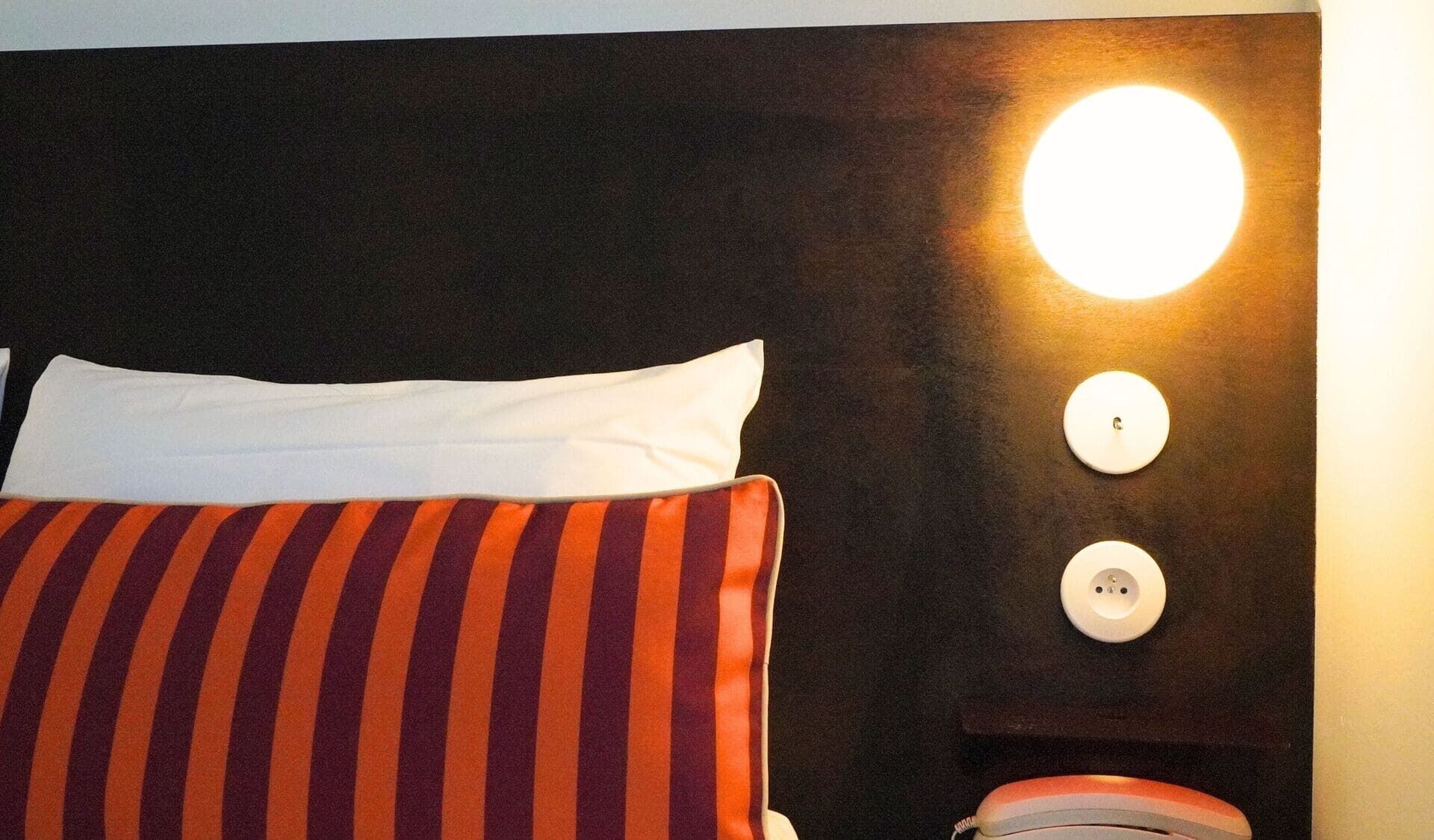 A bed with striped pillows, a white pillow, and a wall-mounted light fixture.