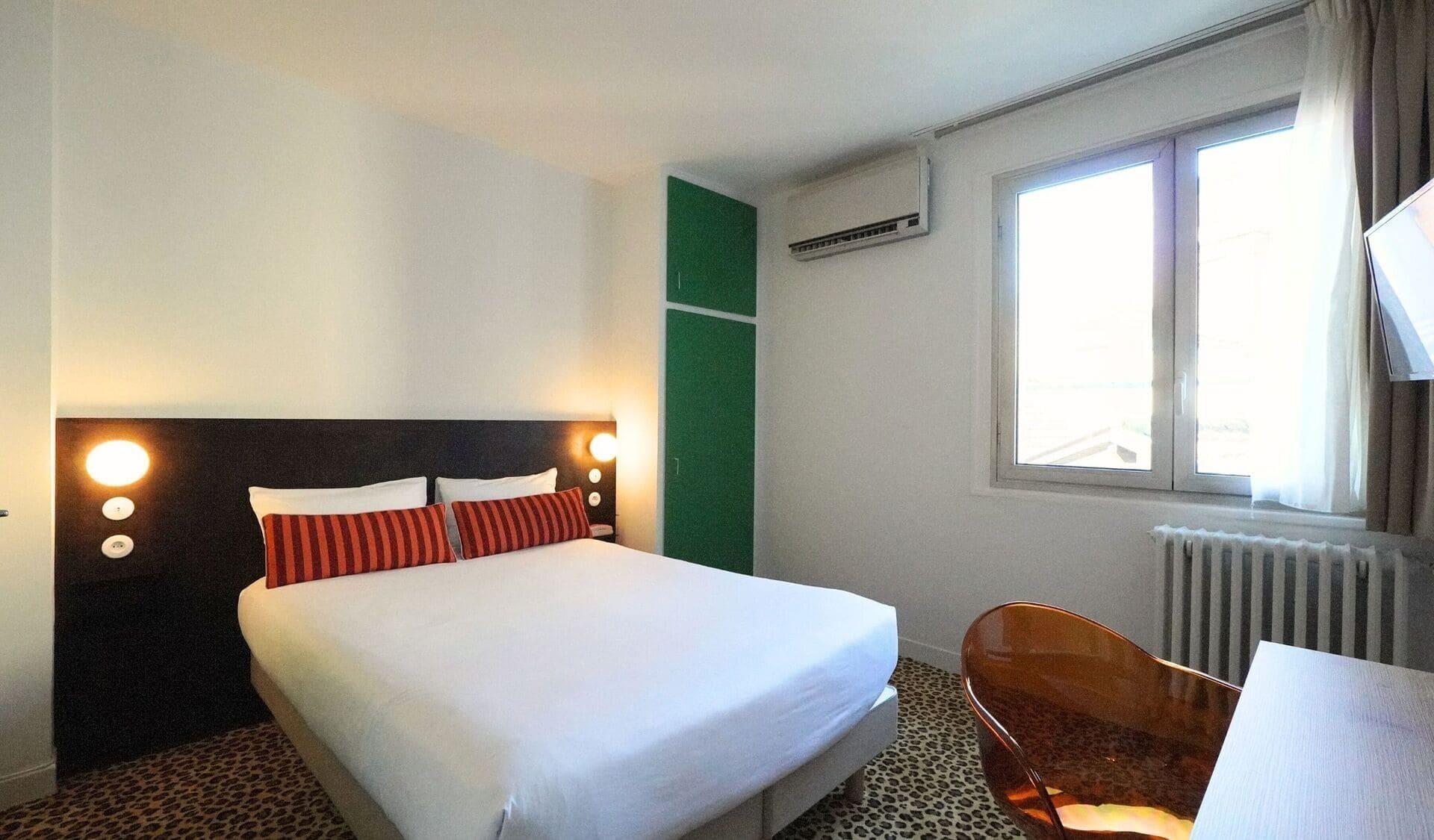 A hotel room with a bed, a chair, a window with curtains, and an air conditioning unit.