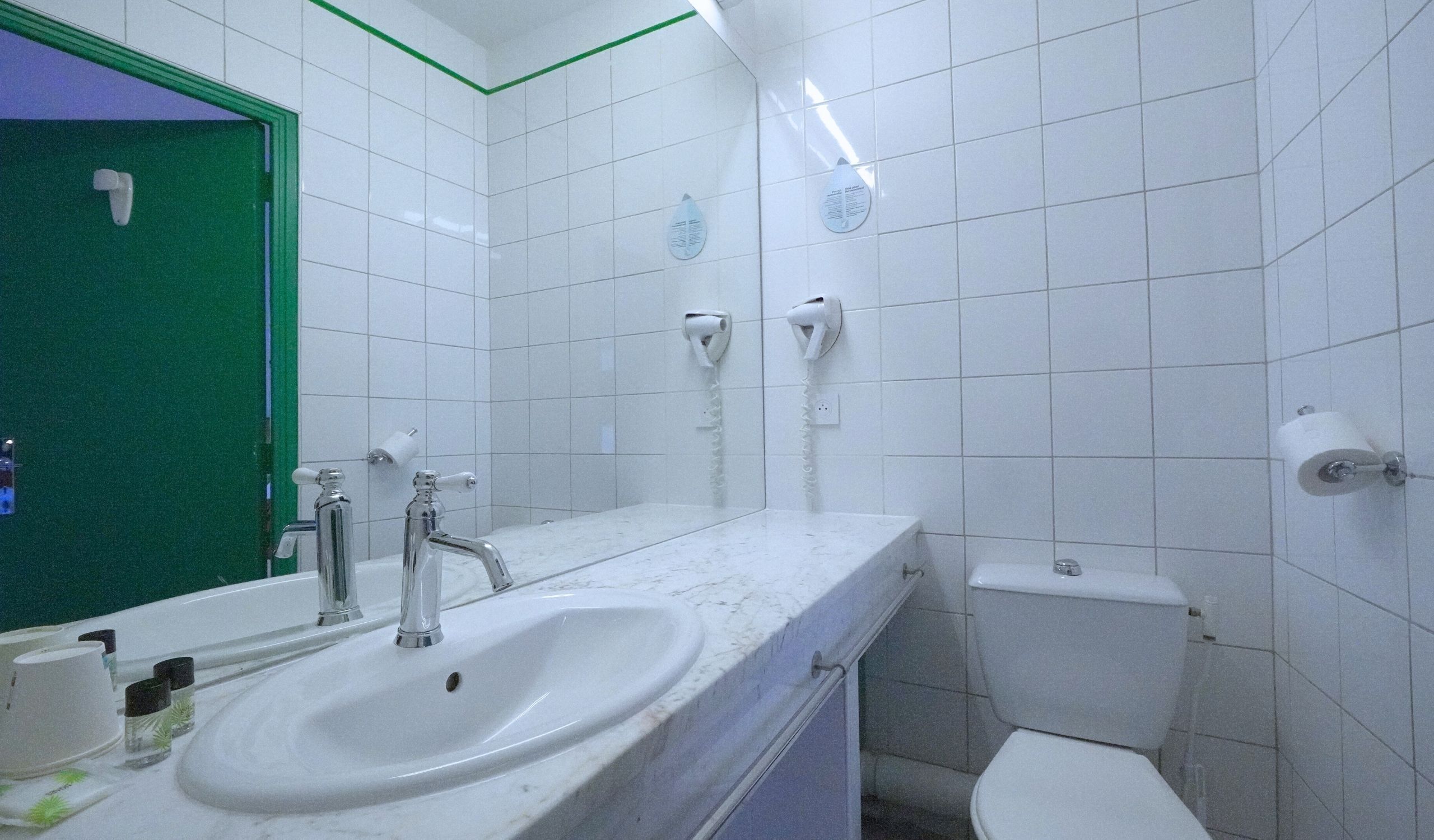 Un baño con un lavamanos blanco, dos secamanos, un inodoro y una puerta verde.