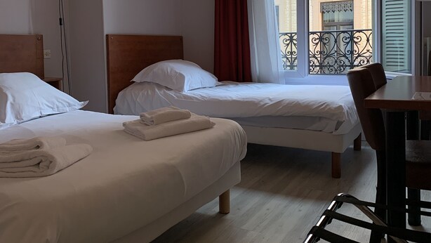 A hotel room with two beds, a desk, a chair, a floor lamp, and a window with a view of buildings. - Wilson Square (Toulouse)