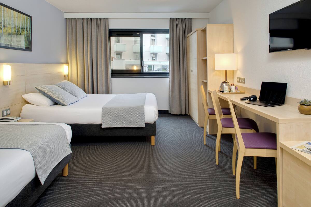 Ein Hotelzimmer mit zwei Betten, einem Schreibtisch mit Laptop, einem Fernseher und einem Fenster mit Vorhängen.
