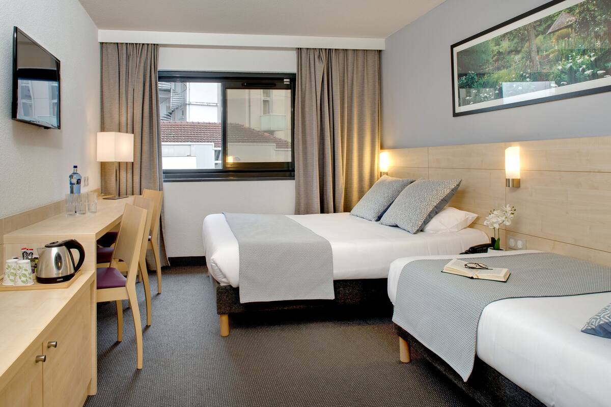 Ein Hotelzimmer mit zwei Betten, einem Schreibtisch, einem Stuhl, einem Fernseher und einem Fenster mit Vorhängen.