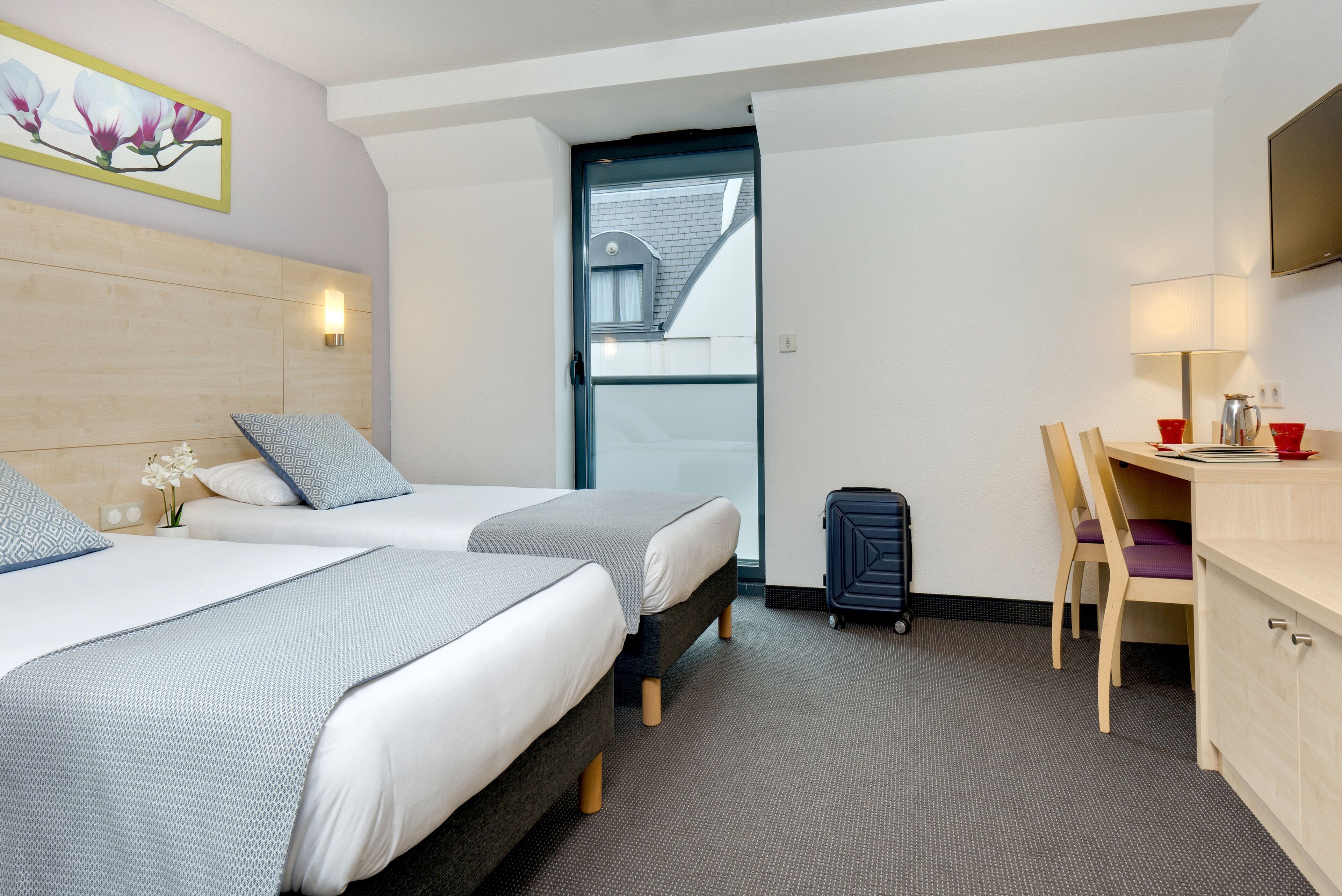 A hotel room with two beds, a desk, a chair, a suitcase, and a window with a view of the outside.