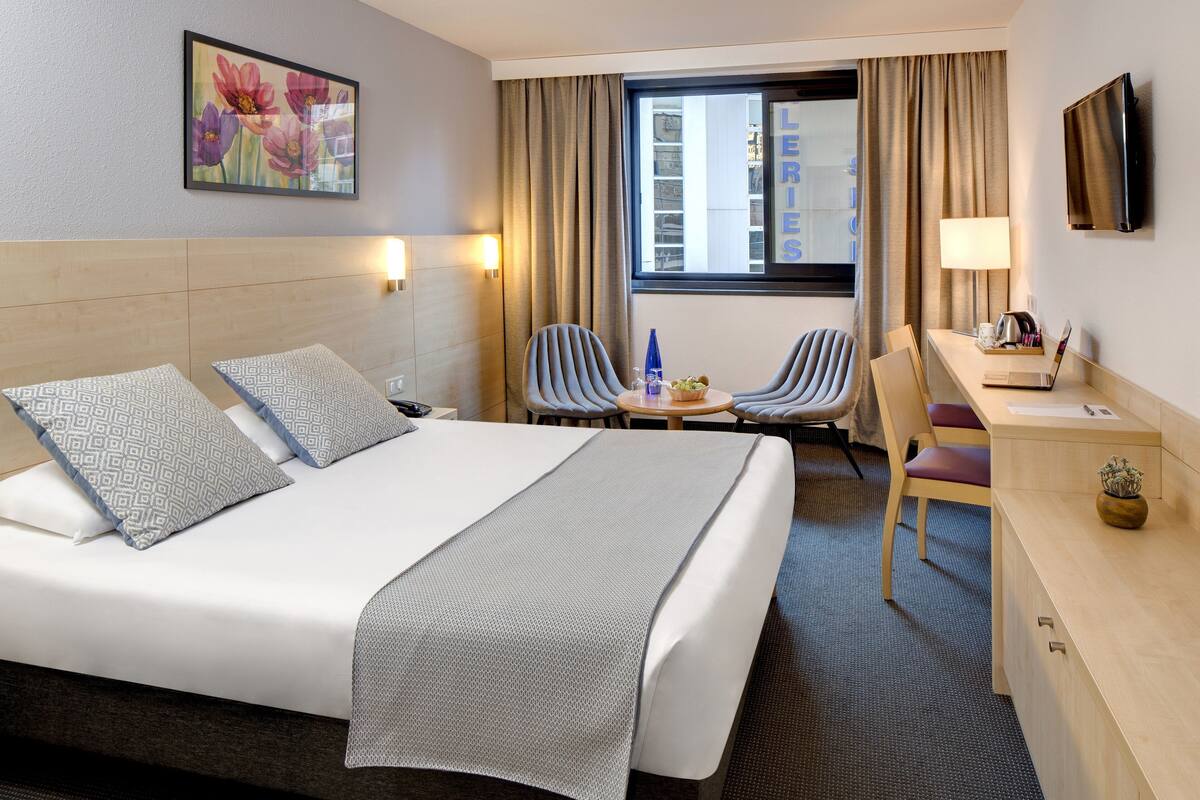 Ein Hotelzimmer mit einem großen Bett, zwei Sesseln, einem Schreibtisch, einem Fernseher und einem Fenster mit Vorhängen.