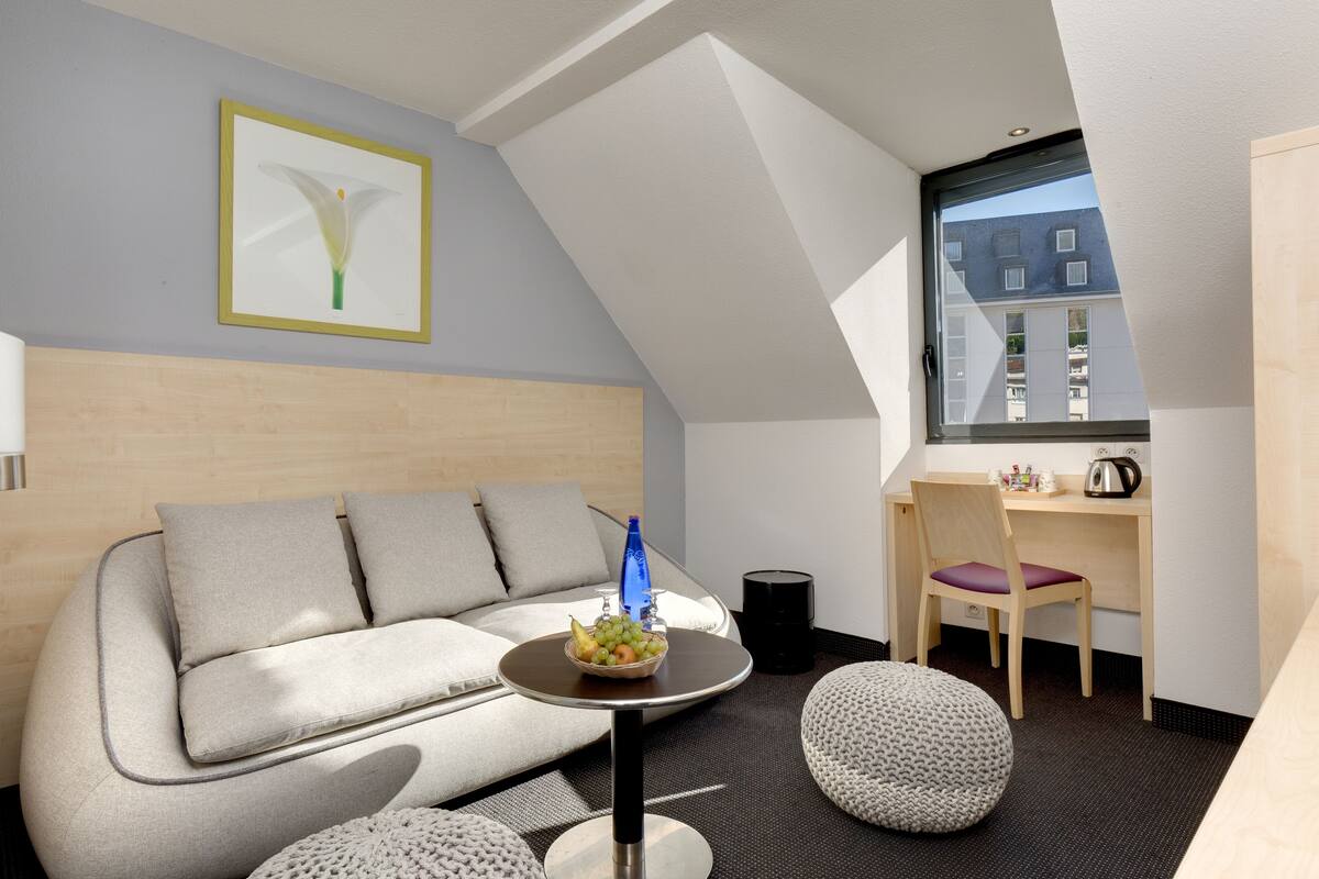 Ein modernes Zimmer mit einer Couch, einem kleinen runden Tisch mit Obstschale, einem Sessel und einem Schreibtisch mit Stuhl.