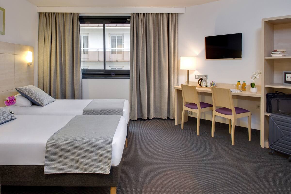Ein Hotelzimmer mit zwei Betten, einem Schreibtisch, einem Stuhl, einem Fernseher und einer Gepäckablage.