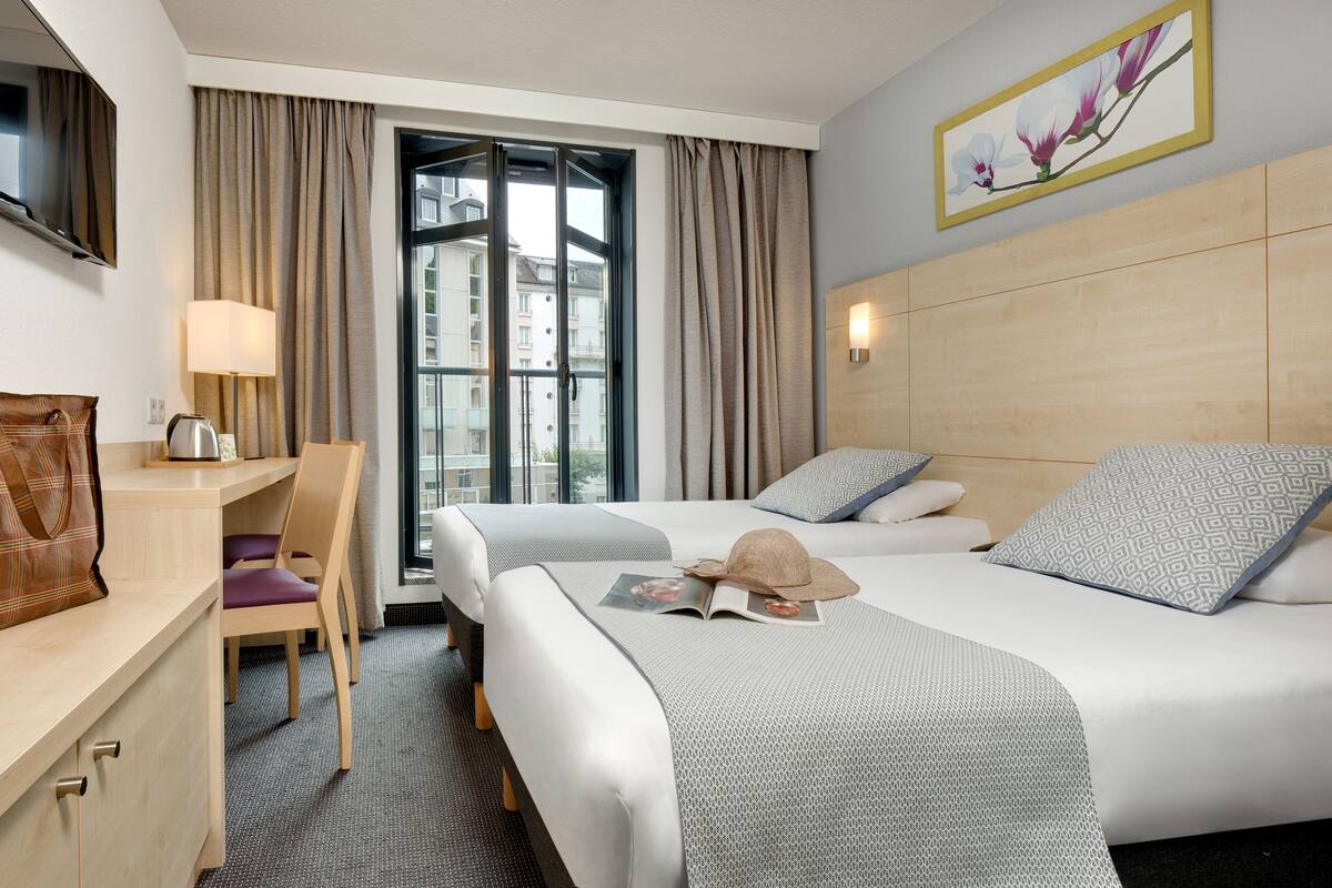 Ein Hotelzimmer mit zwei Betten, einem Schreibtisch, einem Stuhl, einer Lampe und einem Fenster mit Vorhängen.
