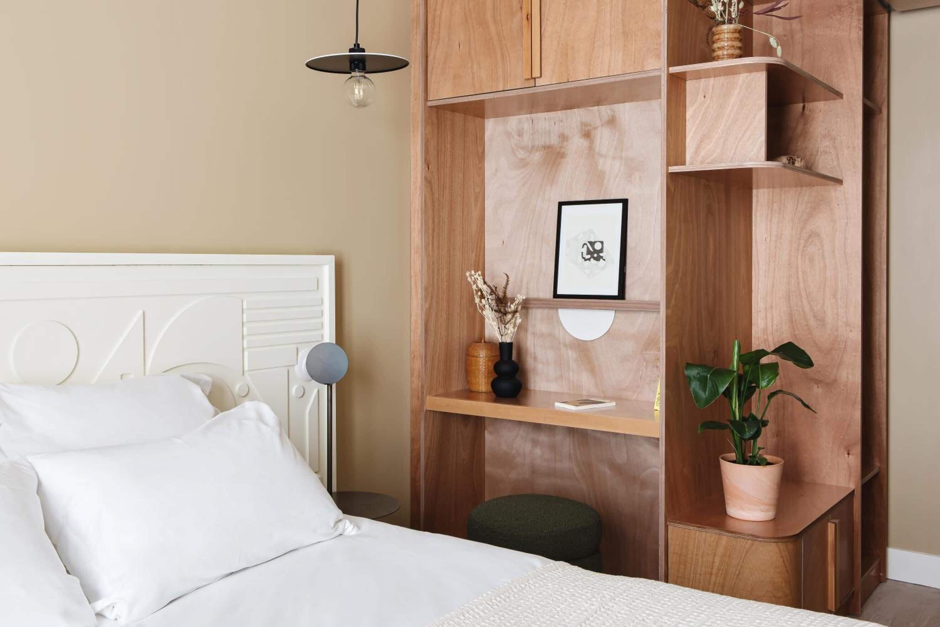 A bedroom with a wooden headboard, a bedside table, a hanging light fixture, a potted plant, and a shelf with decorative items.