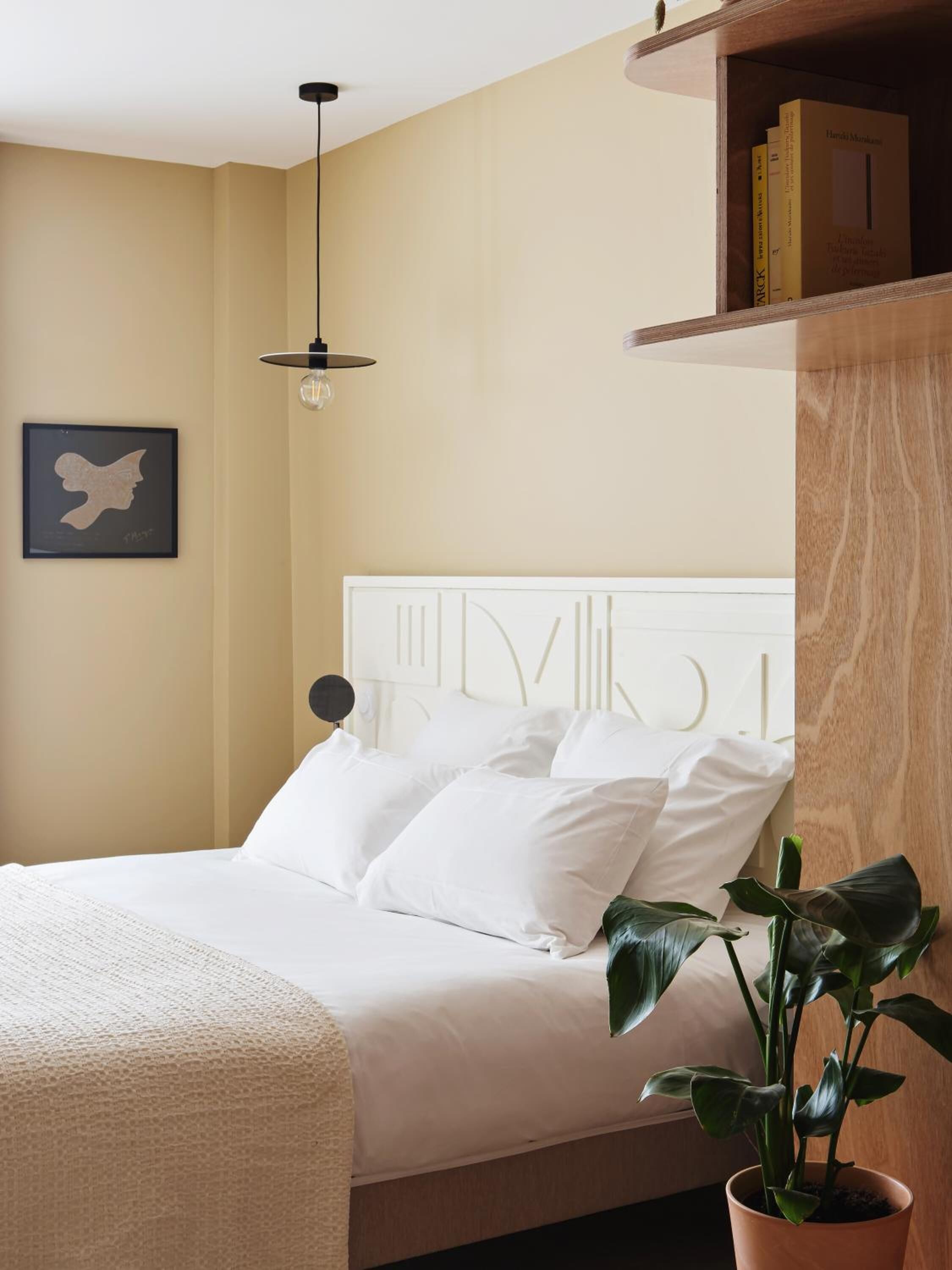 Un lit bien fait, avec une literie et des oreillers blancs, un luminaire suspendu, une œuvre d’art encadrée au mur et une plante en pot posée sur le sol.